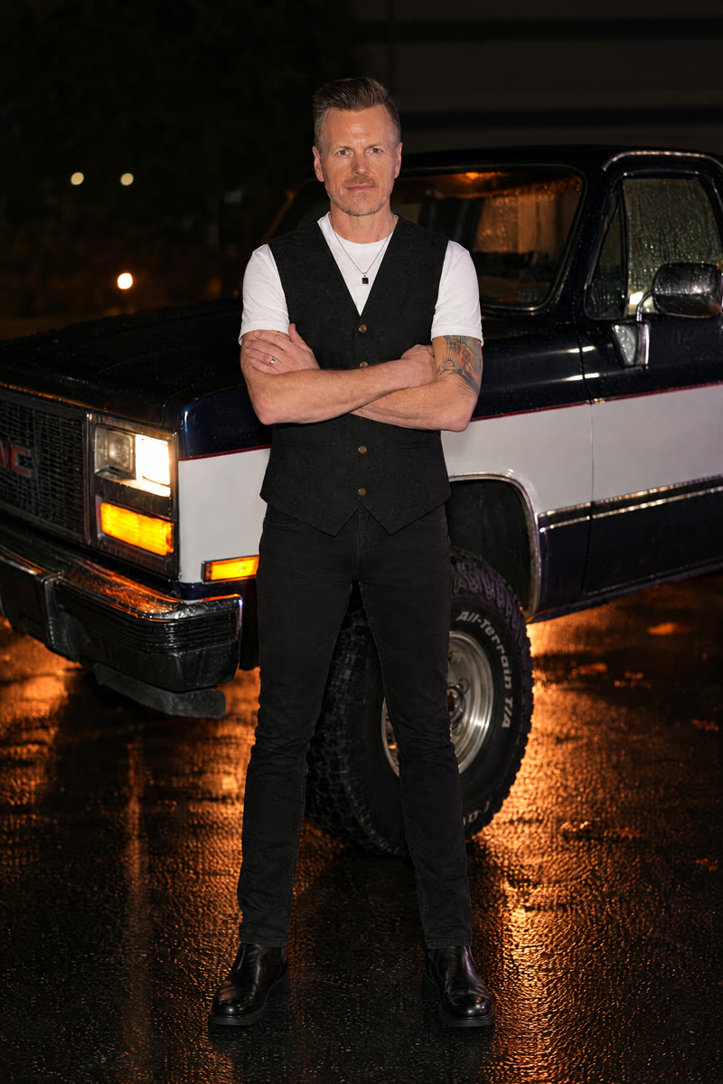 Colin (guitar) in a white T-shirt and black vest, arms crossed in front of the truck.