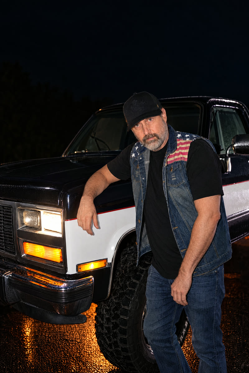 Chris Seibel (drums) in a black cap and flag-patch denim vest, leaning on the truck.